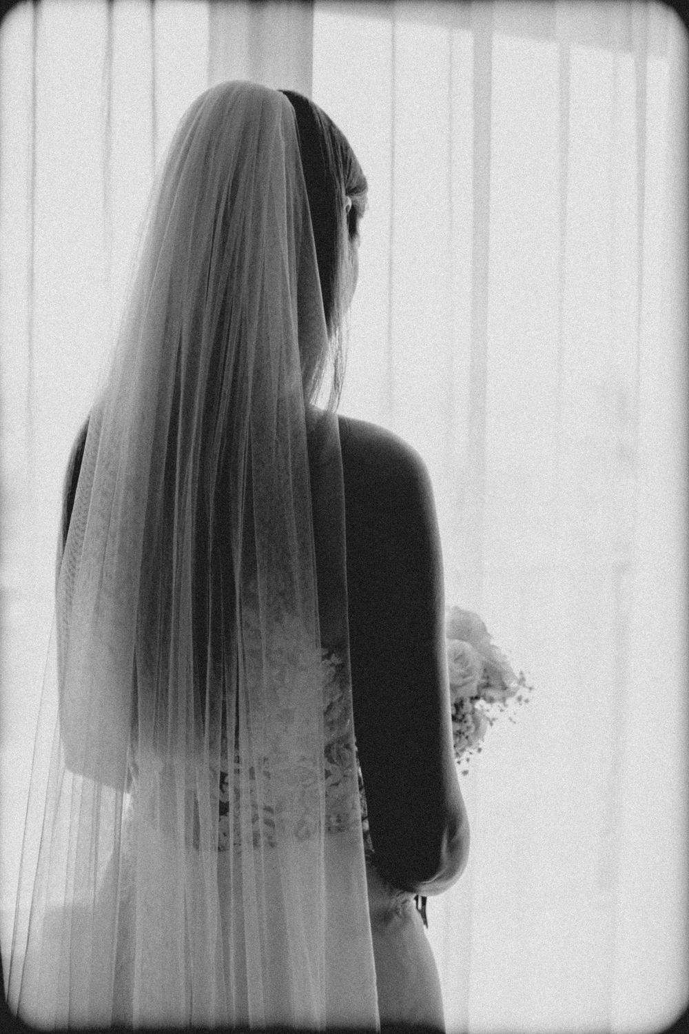 A graceful black and white silhouette of a bride in her veil and wedding dress against sheer white curtains.