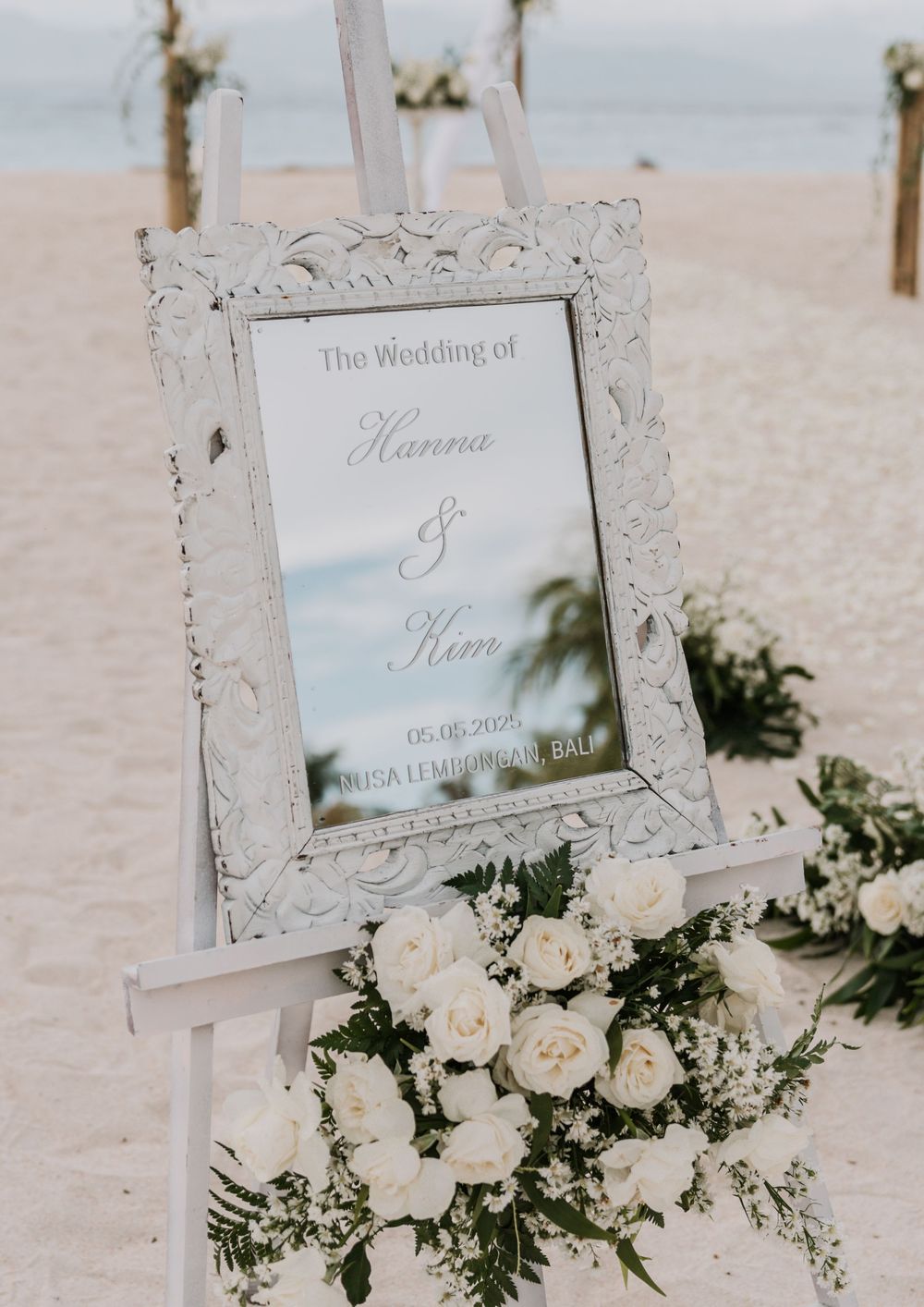 An ornate white frame with a wedding sign sits on an easel at a beach ceremony, surrounded by white roses and greenery.