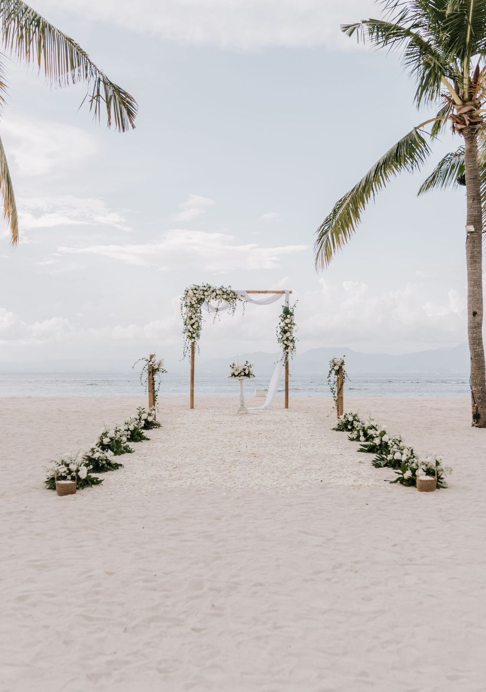 Tropical beach wedding ceremony setup with palm trees and flower arrangements lining a sandy aisle near the ocean.
