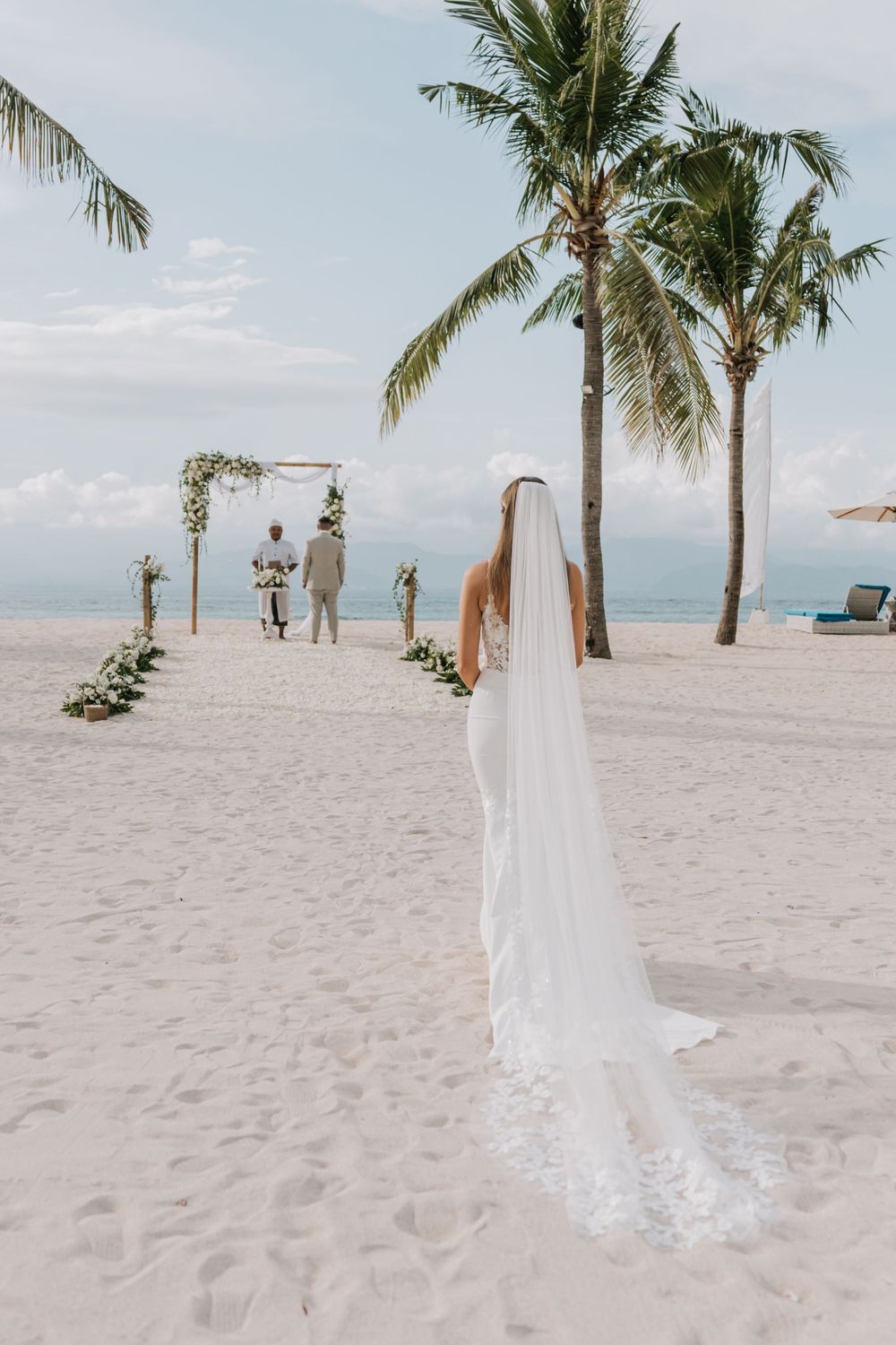 A scenic beach wedding ceremony setup with palm trees, white flowers, and a flowing bridal veil in the foreground.
