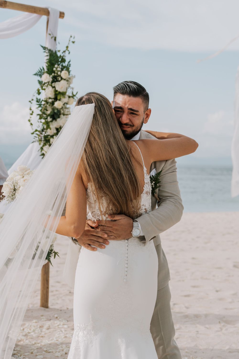 An emotional embrace between a couple during their beachside wedding ceremony with white decor and ocean views.