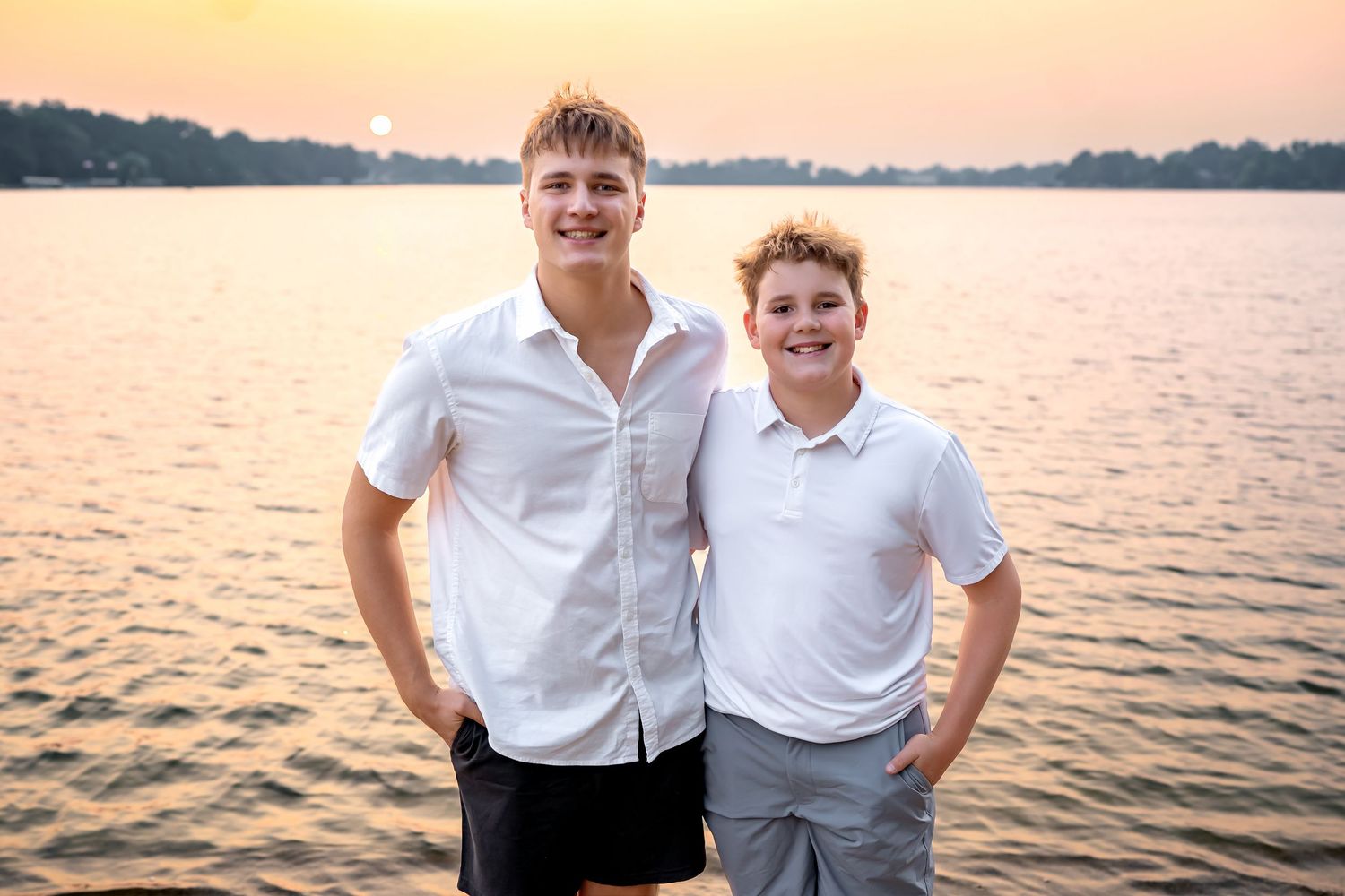 Two people in white shirts pose together at sunset by a peaceful lake with golden light reflecting on the water.