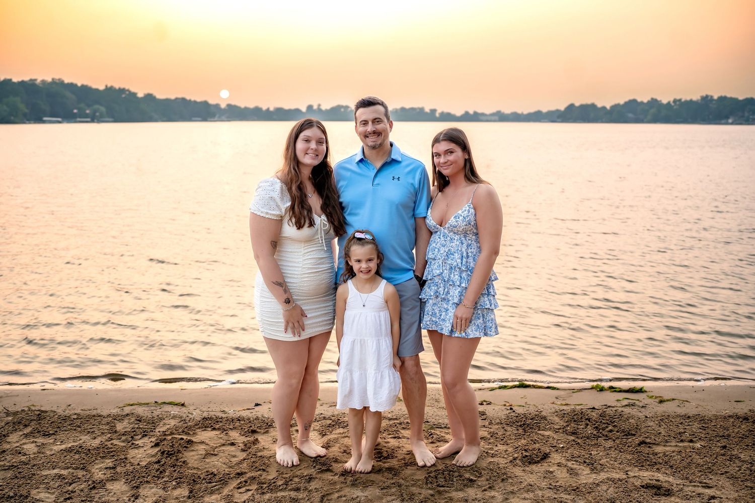A family dressed in coordinating blue and white outfits poses together on the beach during golden hour sunset.