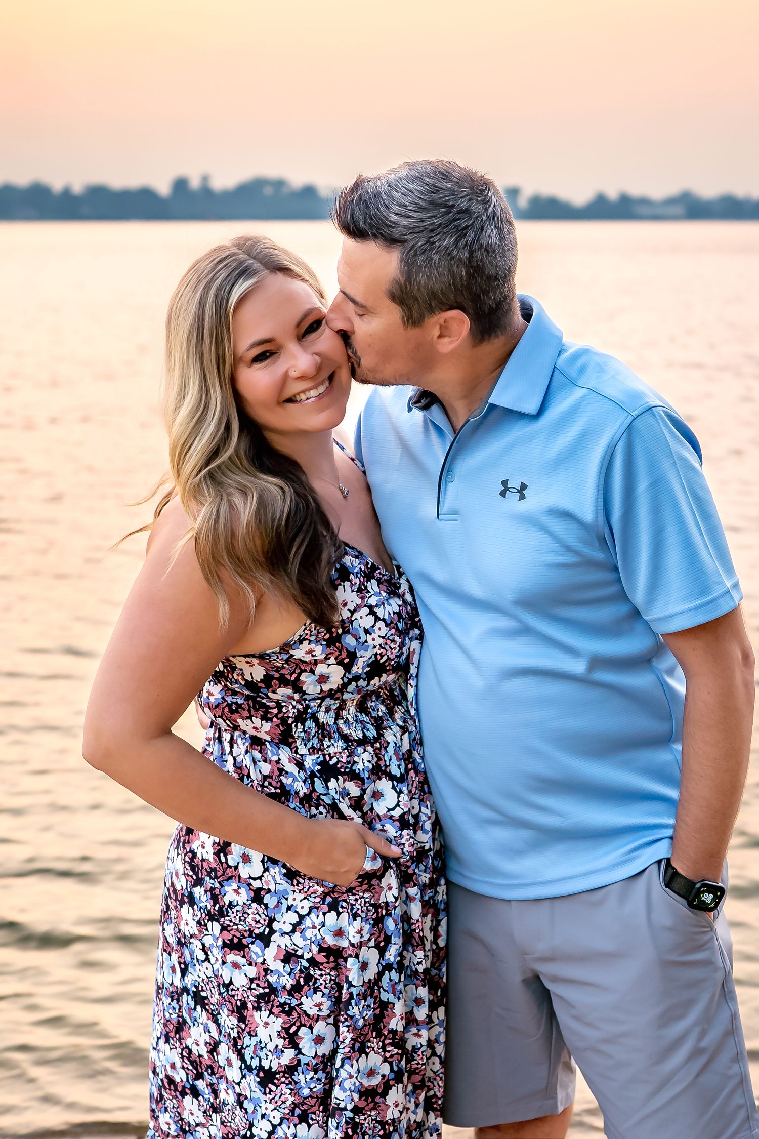 A couple shares a romantic moment on the beach at sunset wearing blue and floral outfits.