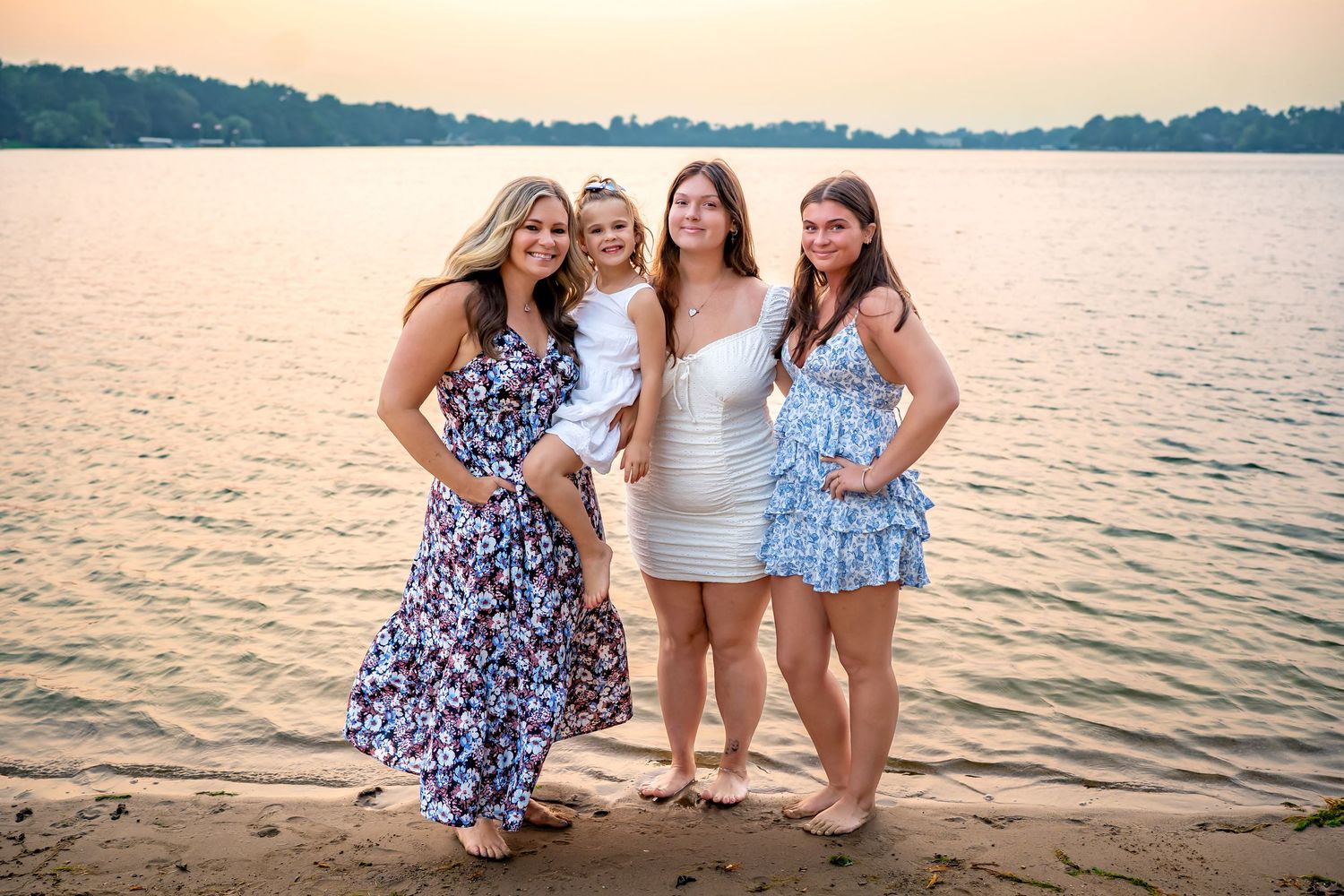 Five friends in sundresses and swimwear pose together at sunset by a tranquil lake.