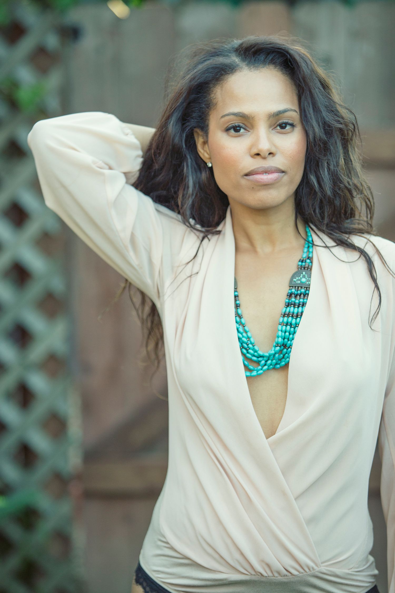 Woman in beige wrap top with turquoise beaded necklace posing outdoors near wooden lattice backdrop.