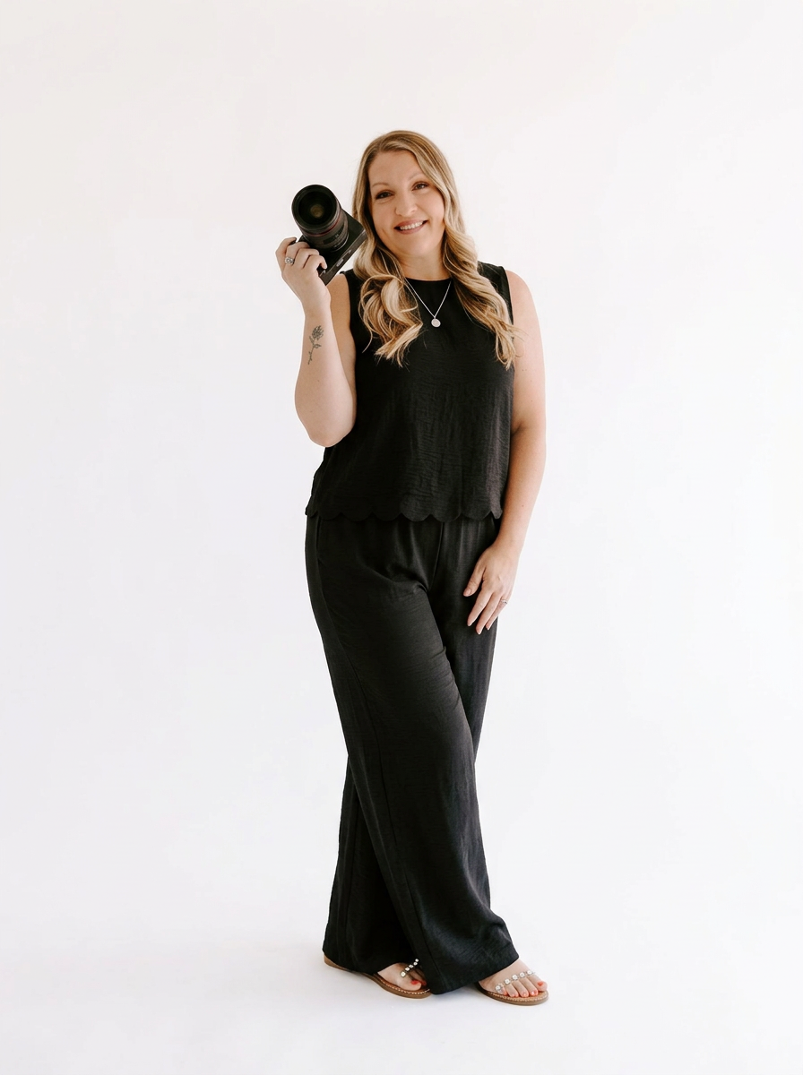 A smiling blonde woman in a black sleeveless jumpsuit holds a camera, posing against a white background.
