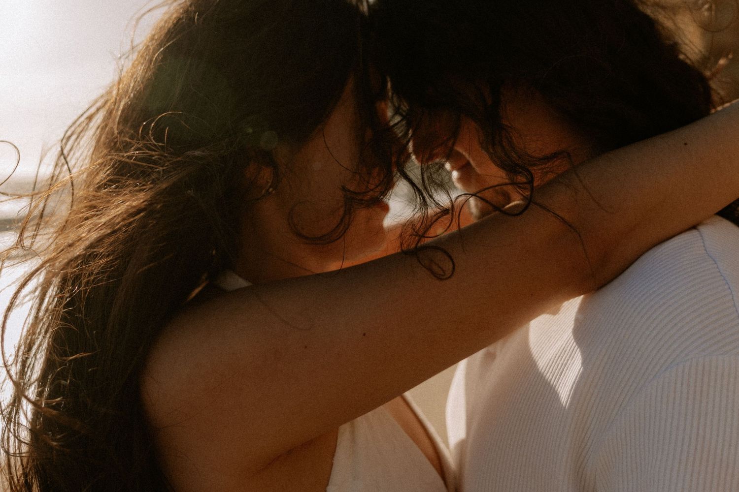 Couple embracing and kissing on a sandy beach with rocky cliffs and ocean waves in the background.