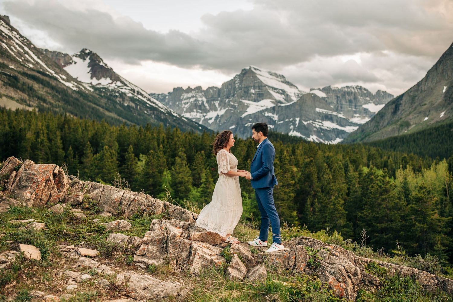 Couple holding hands at mountain elopement with snow-capped peaks and evergreen forest backdrop in Glacier National Park.