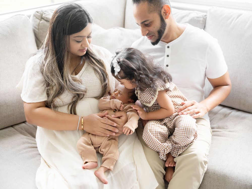 Pregnant woman and man sit on sofa with toddler daughter kissing baby bump in a warm family portrait.
