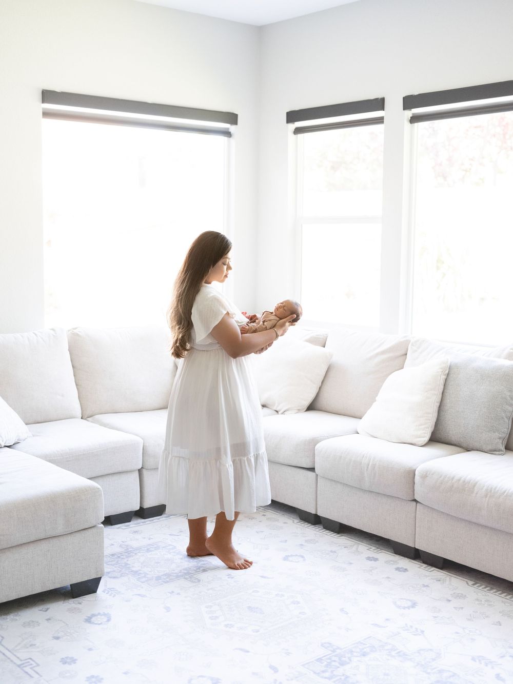 A woman in a white dress holds her newborn baby in a bright, minimalist living room with white sofas.