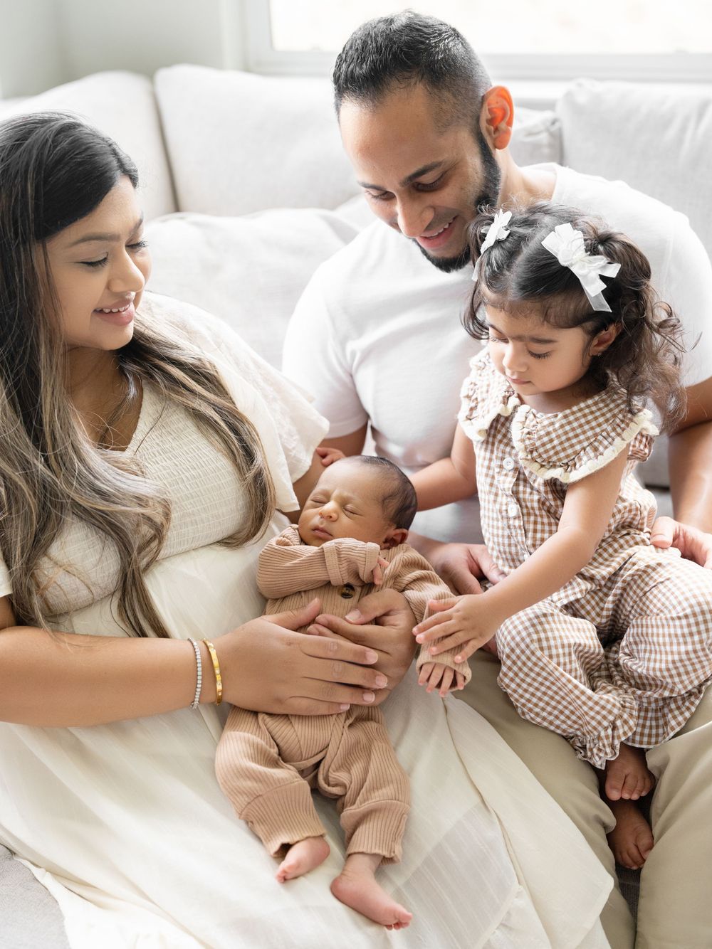 A family of four admires their newborn baby together on a white couch, with a toddler girl in a brown outfit.