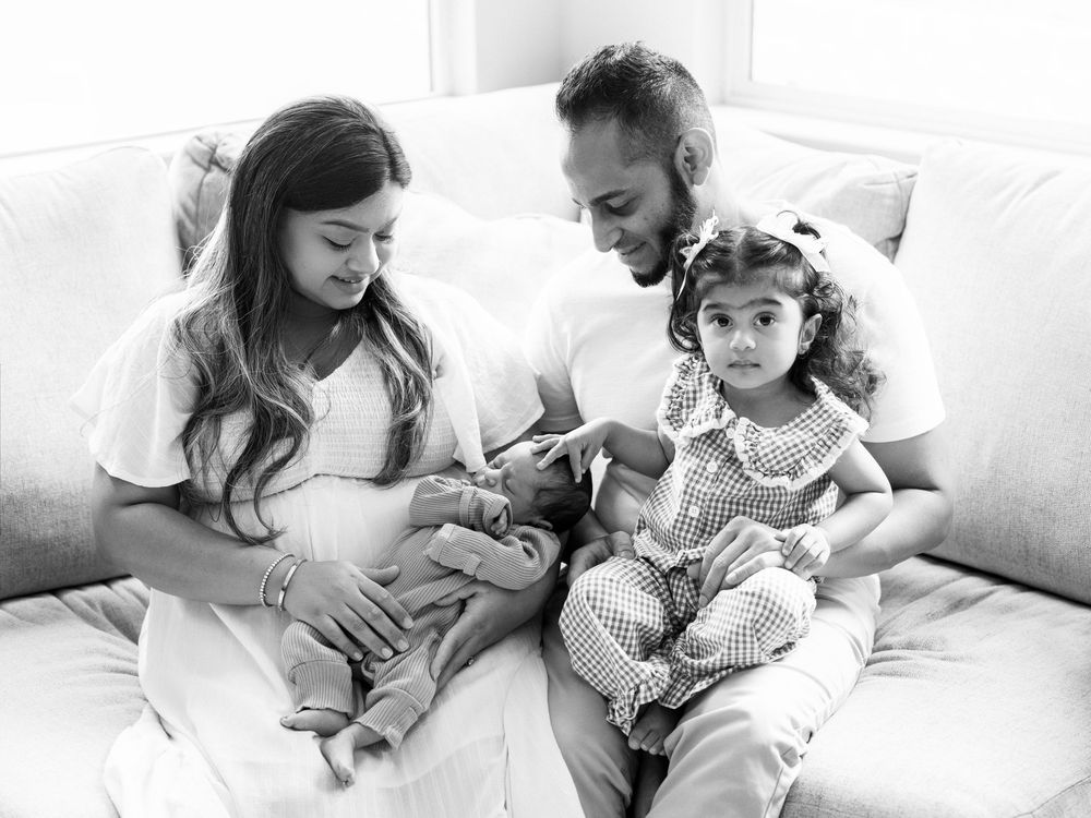 Black and white family portrait of parents with newborn baby and toddler daughter sitting on a couch.