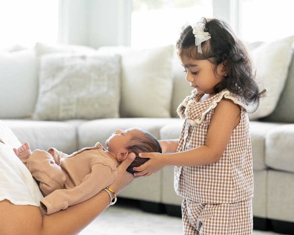 Young girl in brown checkered outfit gently touches newborn baby held by adult in bright, modern living room.