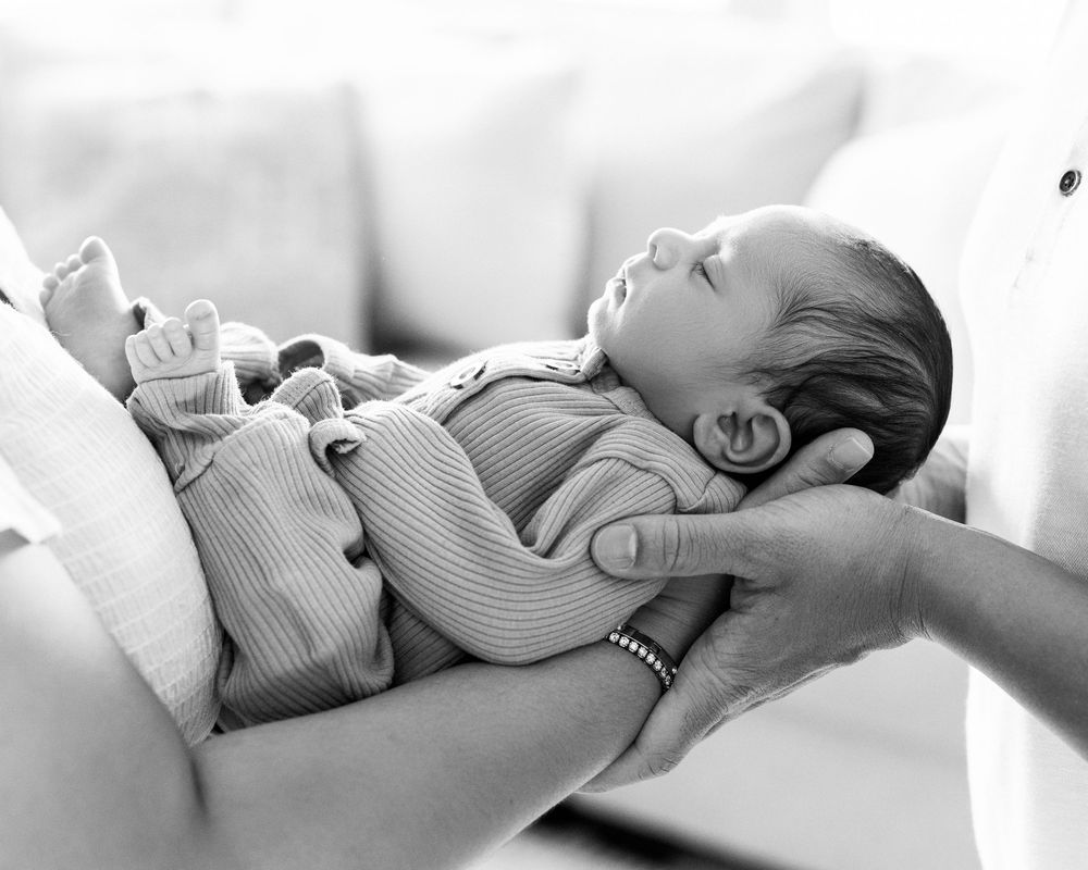 Black and white photo of a sleeping newborn baby cradled gently in adult hands, wearing a ribbed onesie.