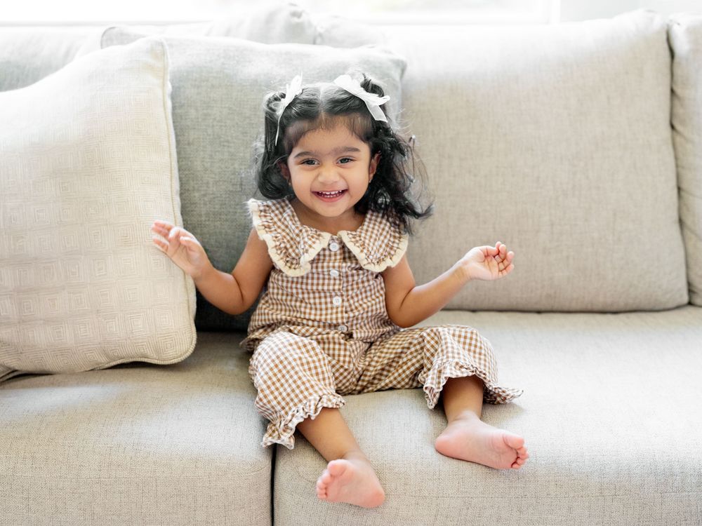 A happy toddler girl in a brown gingham outfit sits smiling on a beige sofa with a white bow in her hair.