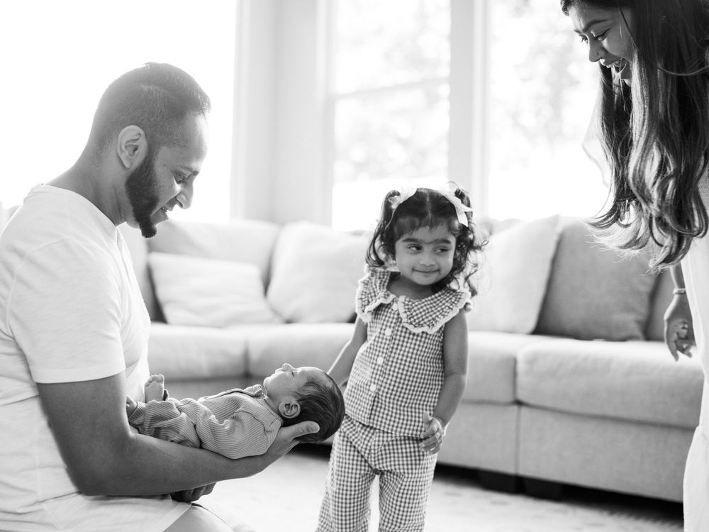 Black and white family photo of father holding newborn baby with toddler daughter and mother in bright living room.