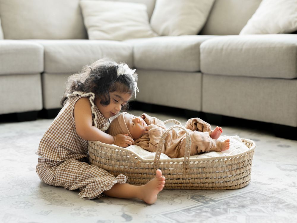 Young toddler girl sitting on floor leaning over a woven Moses basket with a newborn baby inside.