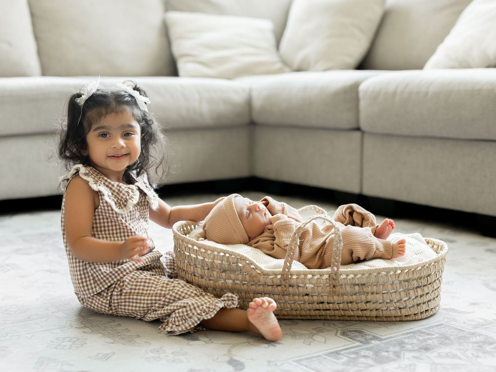 Toddler girl in gingham dress sits beside a newborn baby in a wicker Moses basket on a living room floor.