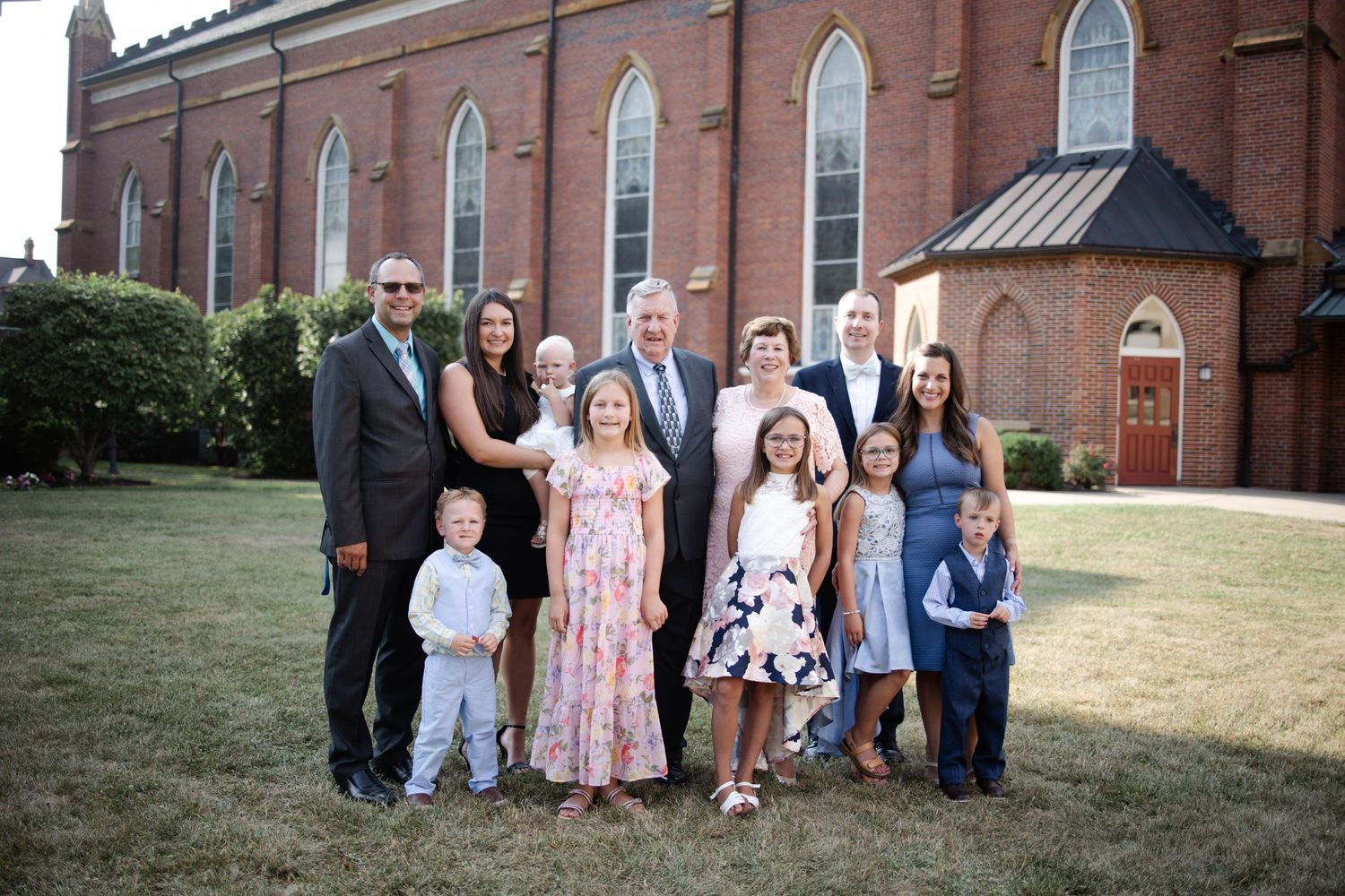 Family portrait of multi-generational group posing outdoors in front of a red brick church on a sunny day.