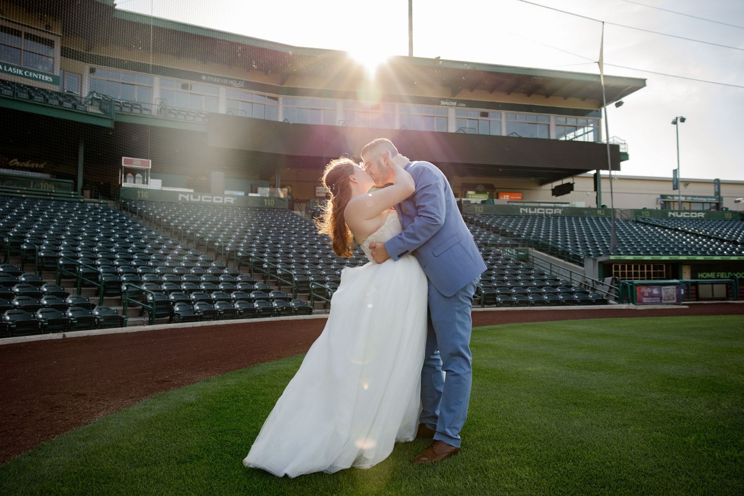 Bride and groom share romantic kiss on baseball stadium field at golden sunset hour.