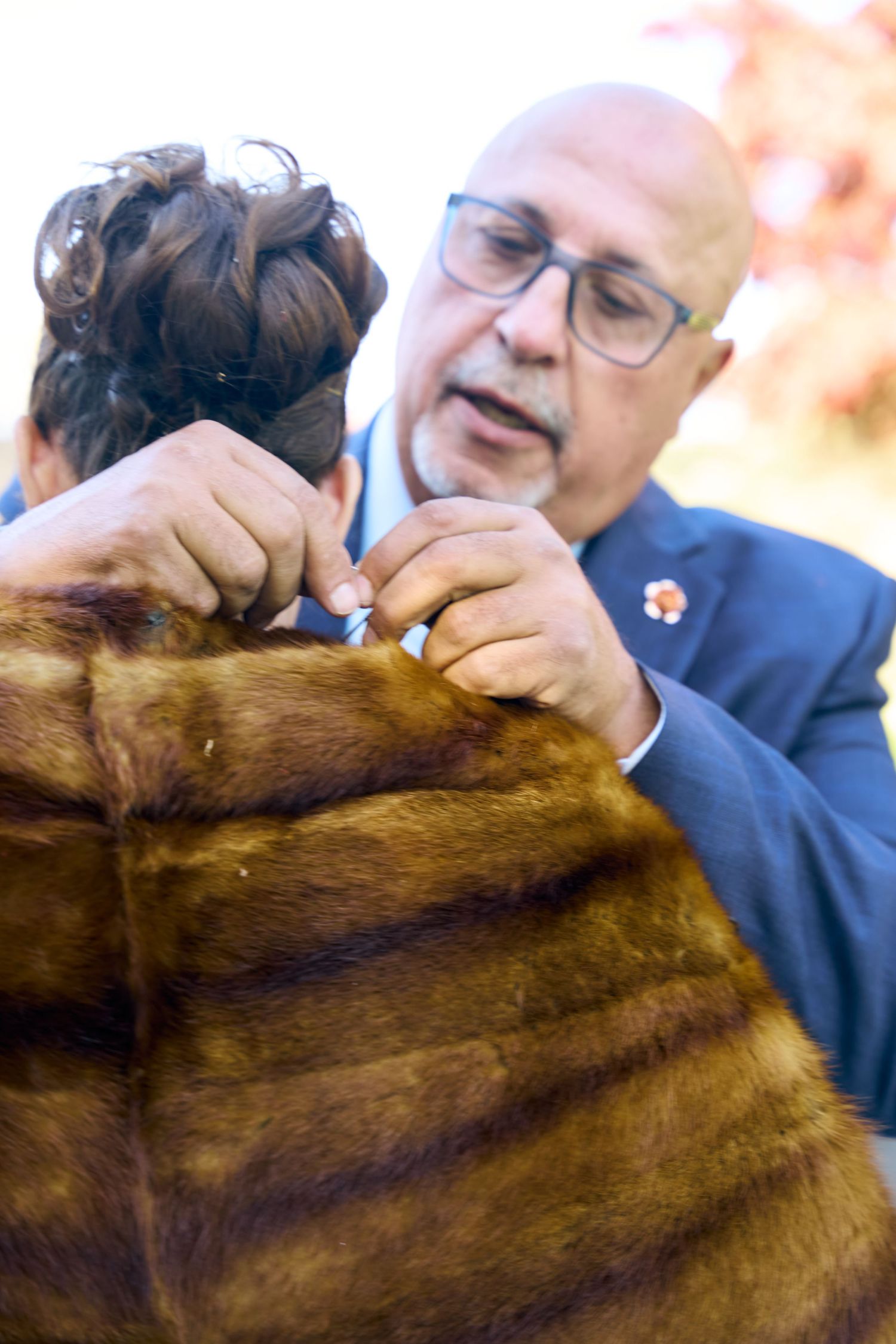 A bald man with glasses fastens a fur coat on a woman with an elegant updo hairstyle outdoors.
