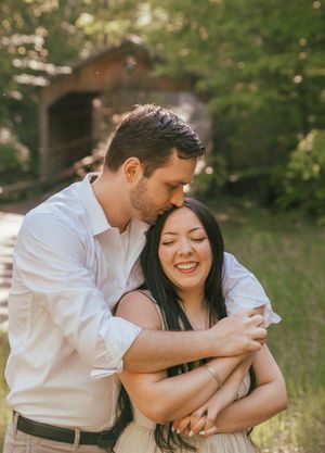 Couple embracing outdoors near a covered bridge, man kissing woman's head while she smiles joyfully.