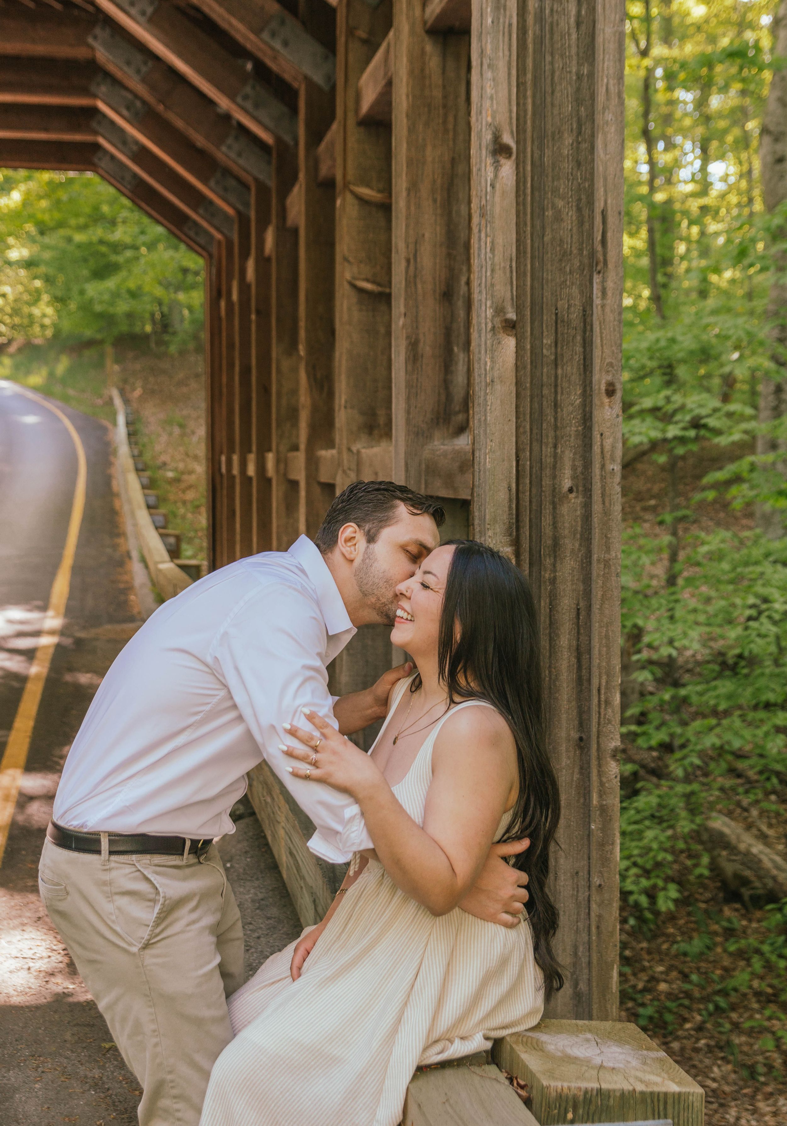 Couple shares a romantic kiss under a rustic wooden covered bridge surrounded by lush green trees.
