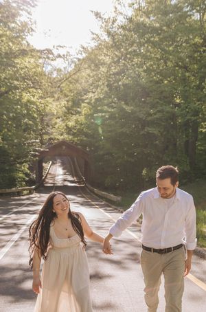A couple walks hand-in-hand on a sunlit road lined with trees, smiling joyfully with a covered bridge in the background.