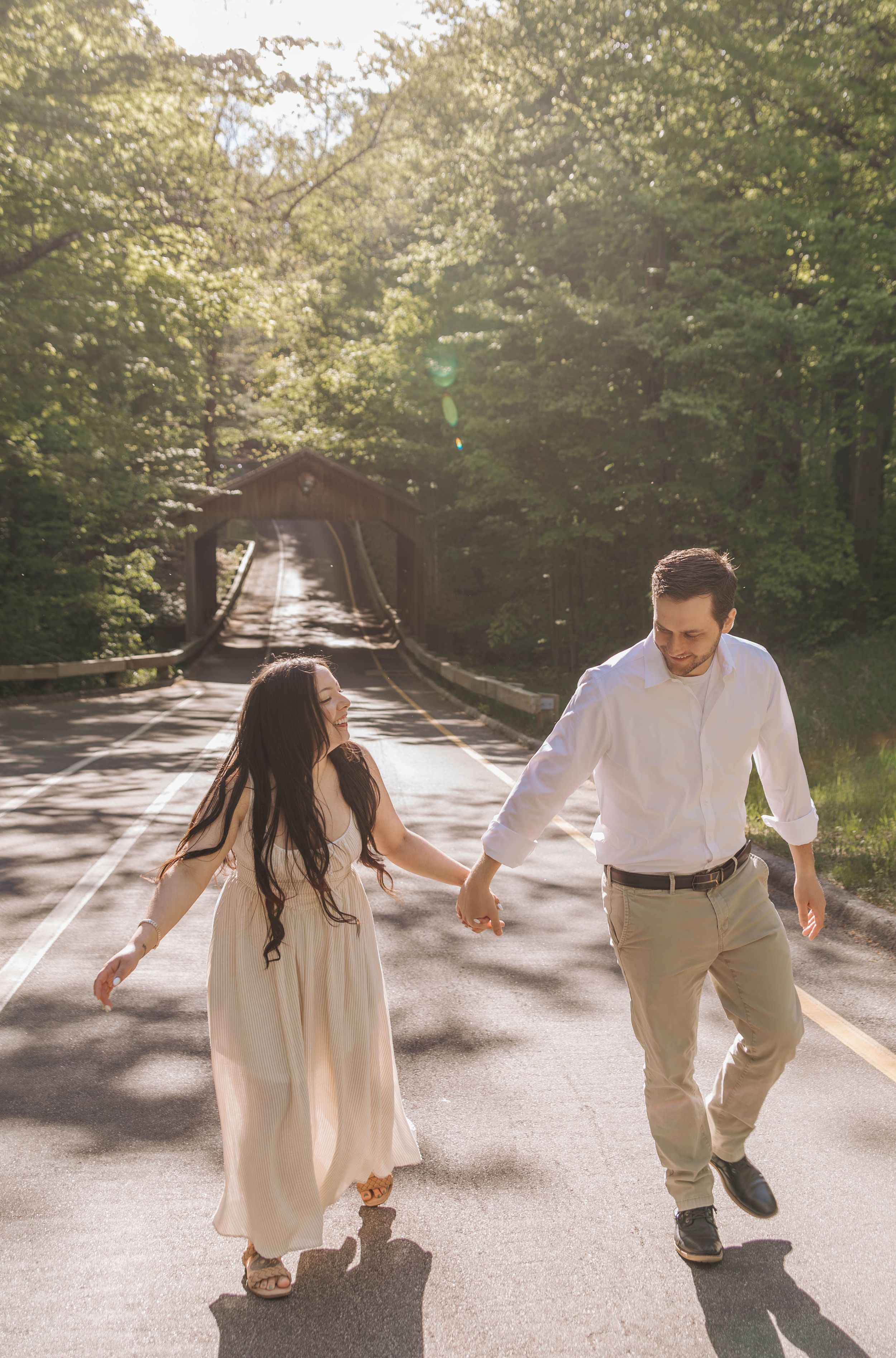 Couple holding hands walking on a sunlit road toward a covered bridge surrounded by lush green trees.