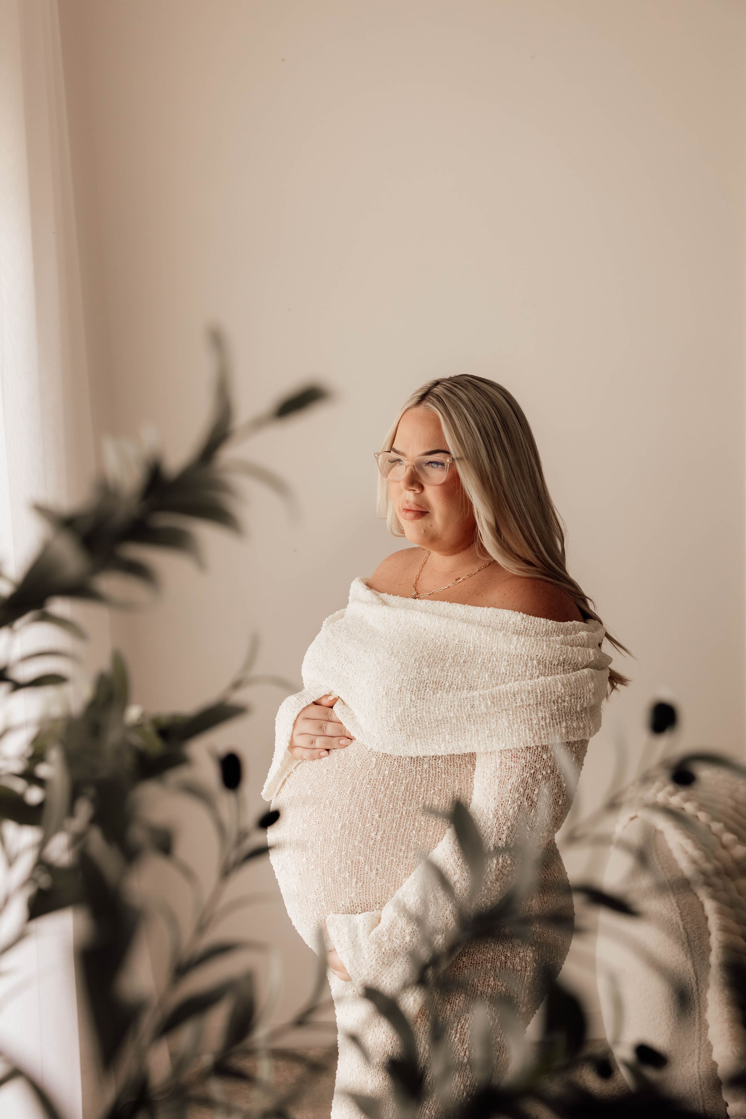 Pregnant woman in white off-shoulder wrap dress cradling baby bump, standing near plants in soft natural light.