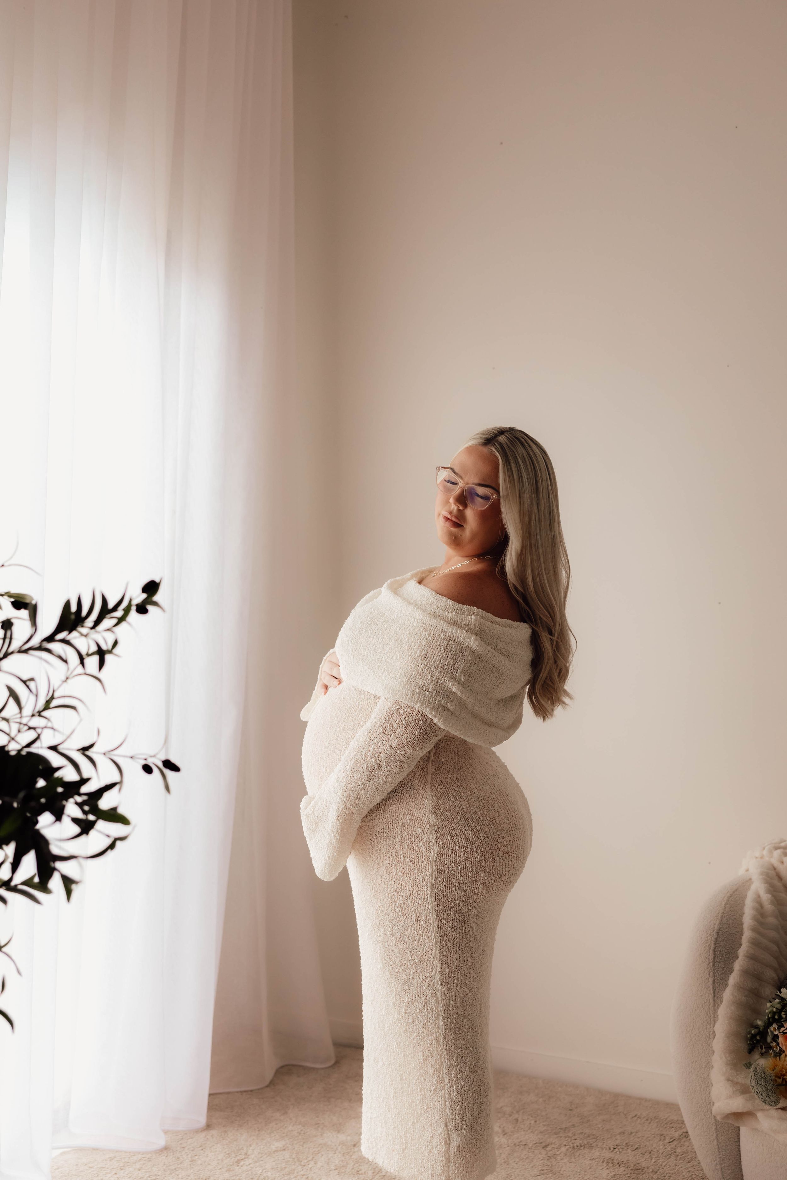 Pregnant woman in white off-shoulder knit dress cradling baby bump near sheer curtains.