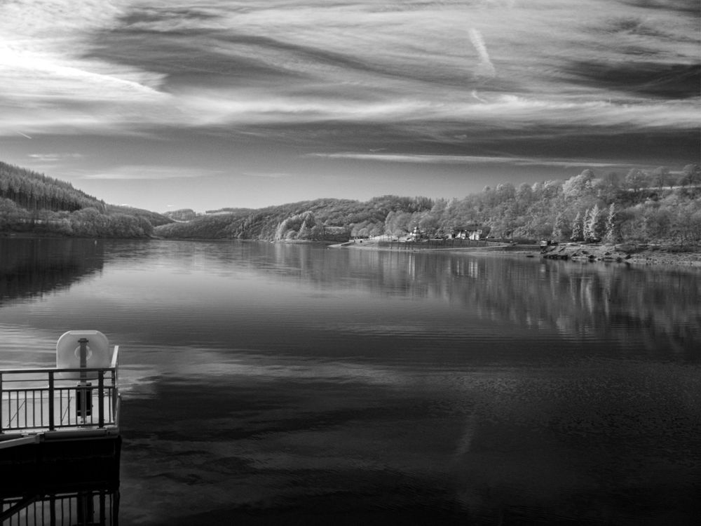 Black and white infrared photo of a calm lake reflecting clouds and forested hills with a dock in the foreground.