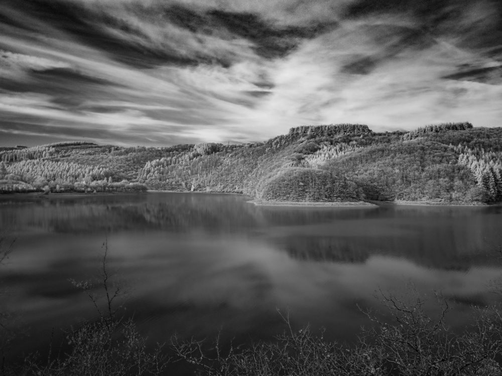 Black and white long exposure photo of a large lake reflecting dramatic wispy clouds with forested hills beyond.