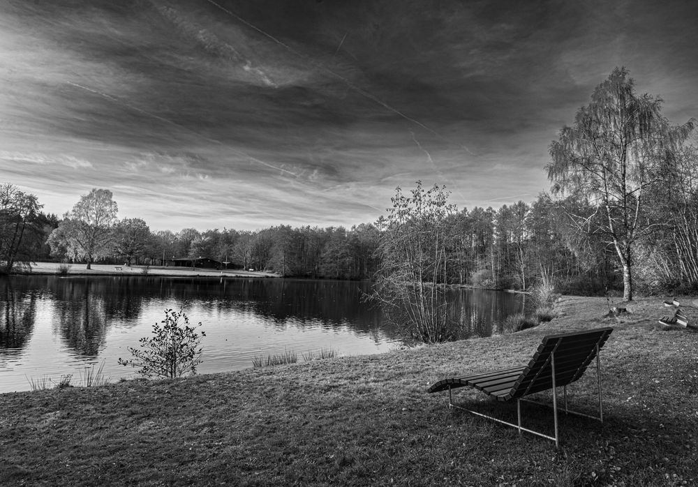 Black and white photo of a lounge chair beside a tranquil lake surrounded by birch trees under a cloudy sky.