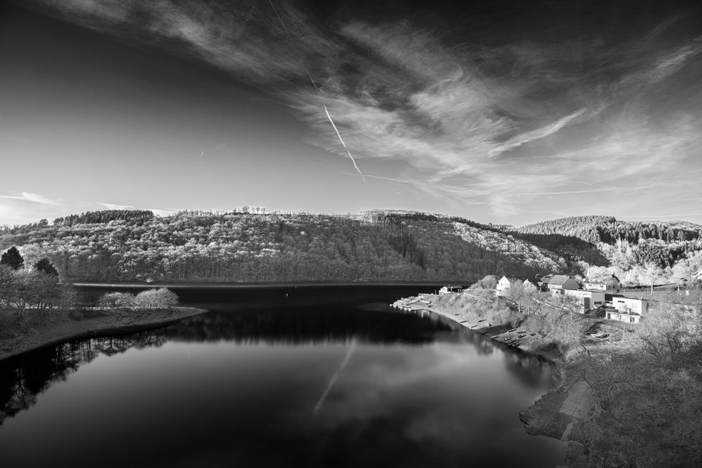 Black and white panoramic photo of a calm reservoir reflecting the sky with forested hills and small buildings.