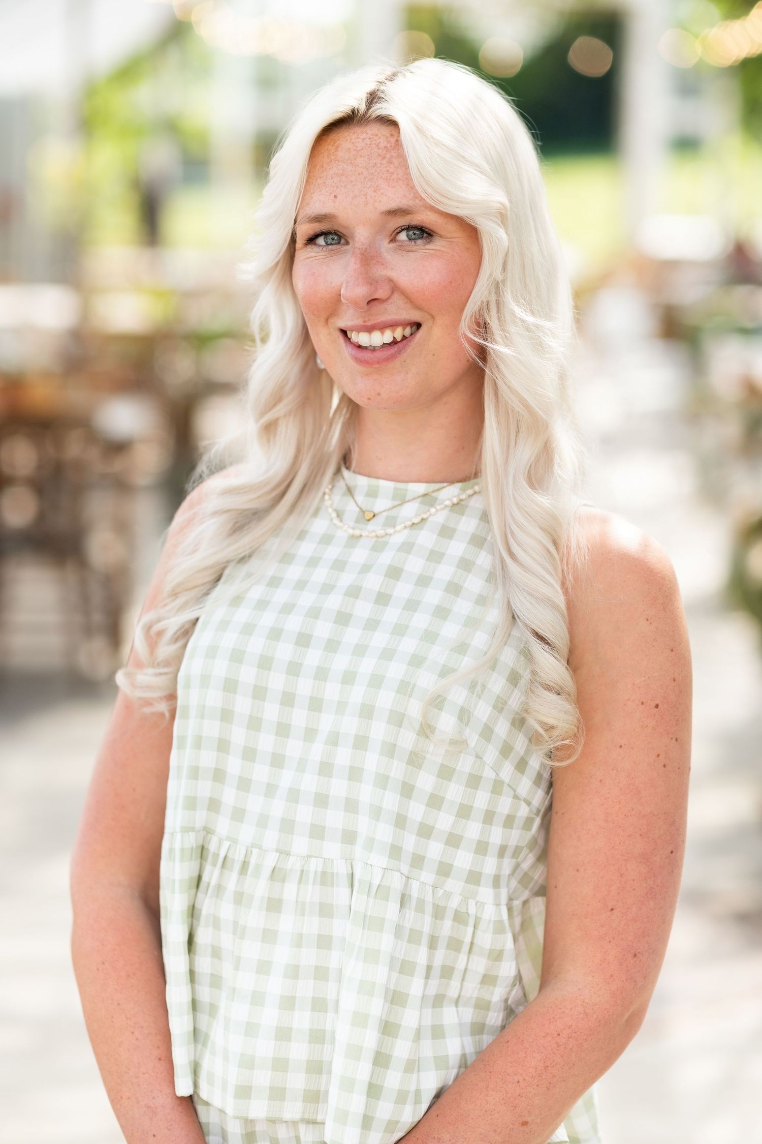 Smiling woman with long blonde hair wearing a green gingham sleeveless dress outdoors.