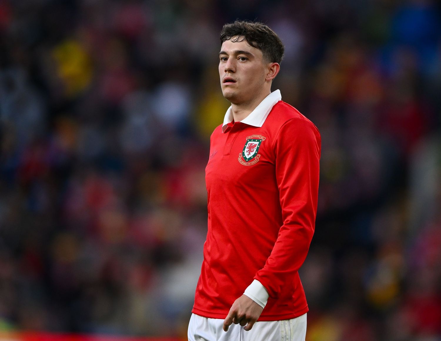 Young male footballer in red Wales national team jersey standing on pitch during a match.