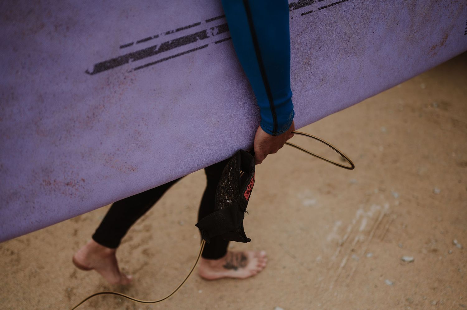 Two surfers in wetsuits carry a purple surfboard on a sandy beach, with a surf leash visible near their bare feet.
