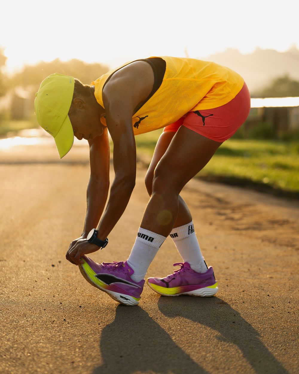 A runner in Puma gear ties their purple and yellow running shoes on a sunlit road.
