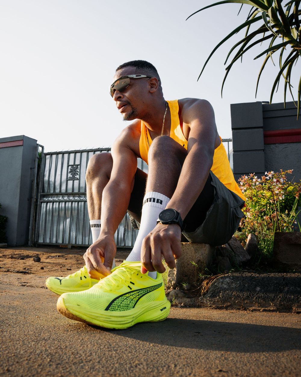 A man wearing Puma neon yellow sneakers and orange vest crouches on a sunny urban street.