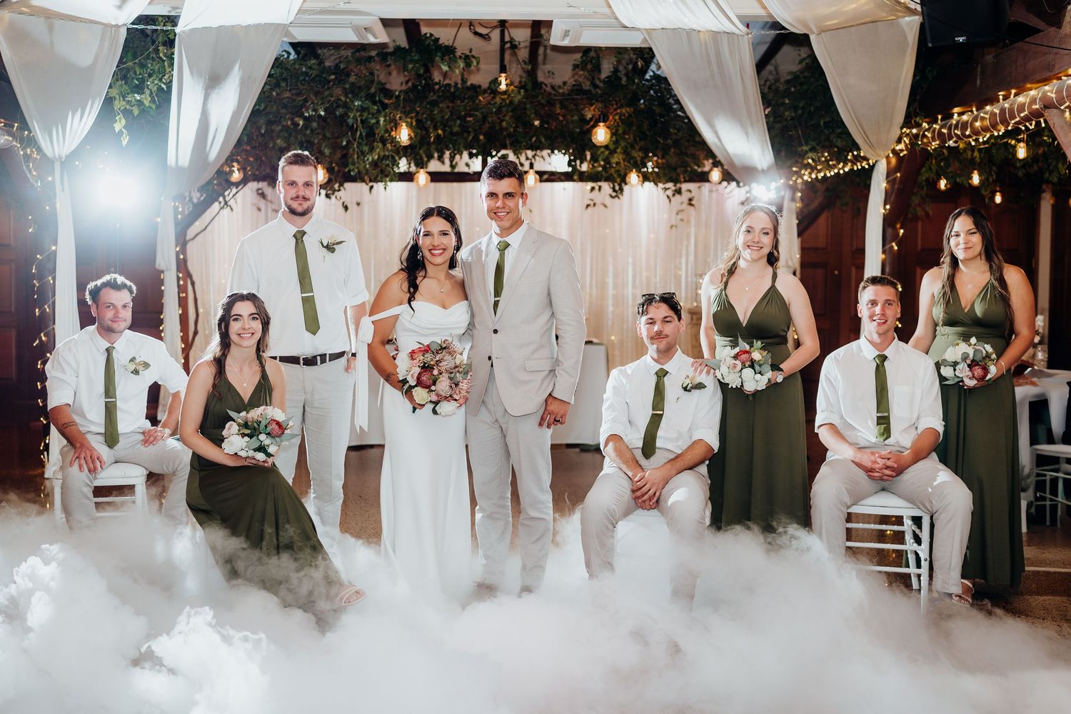 Wedding party poses together on a foggy dance floor, with bridesmaids in green dresses and groomsmen in white suits.