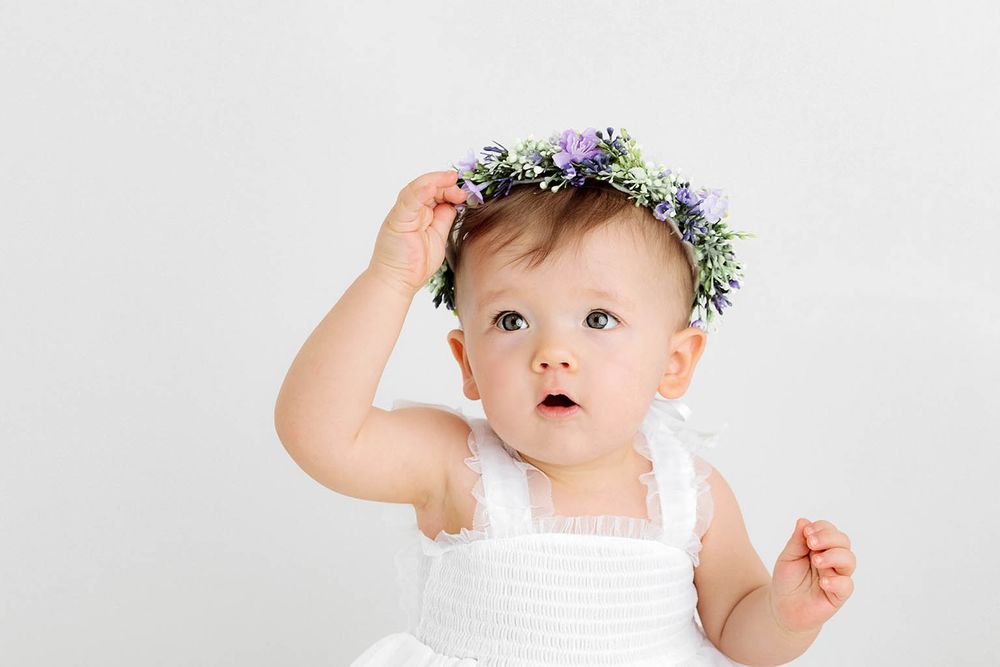 Adorable baby girl in white dress adjusts purple floral crown on her head against white background.