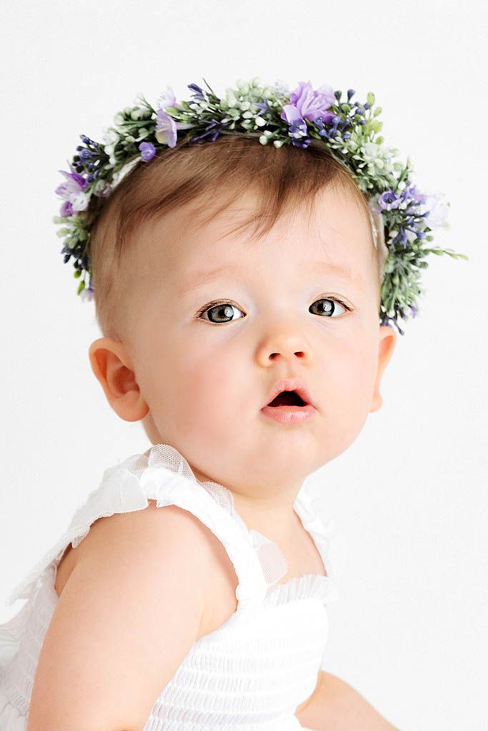 Close-up portrait of baby girl wearing purple and green floral crown in white dress.