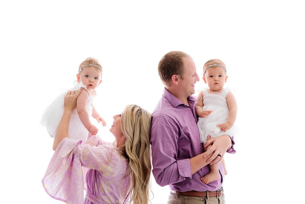 Happy family with parents holding twin baby girls in white dresses against white background.
