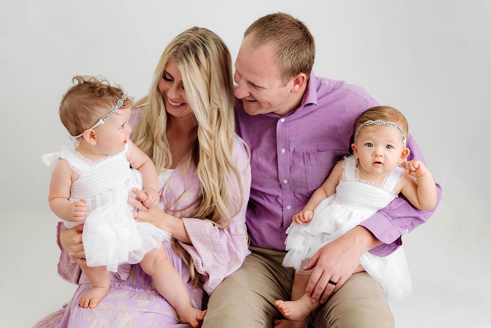 Smiling parents in purple outfits sitting and holding their twin baby girls in white dresses.
