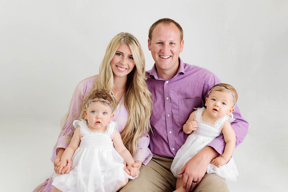 A smiling family of four with two baby girls in white dresses posing together in a bright studio portrait.