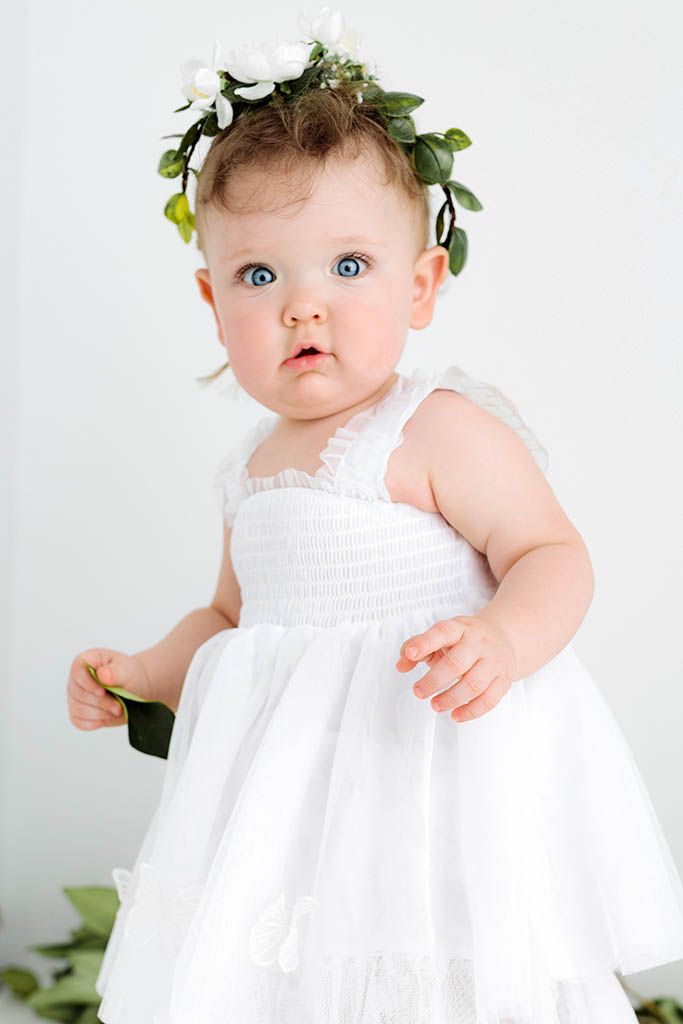 Adorable baby girl in white smocked dress and floral crown with blue eyes, perfect flower girl outfit for weddings.
