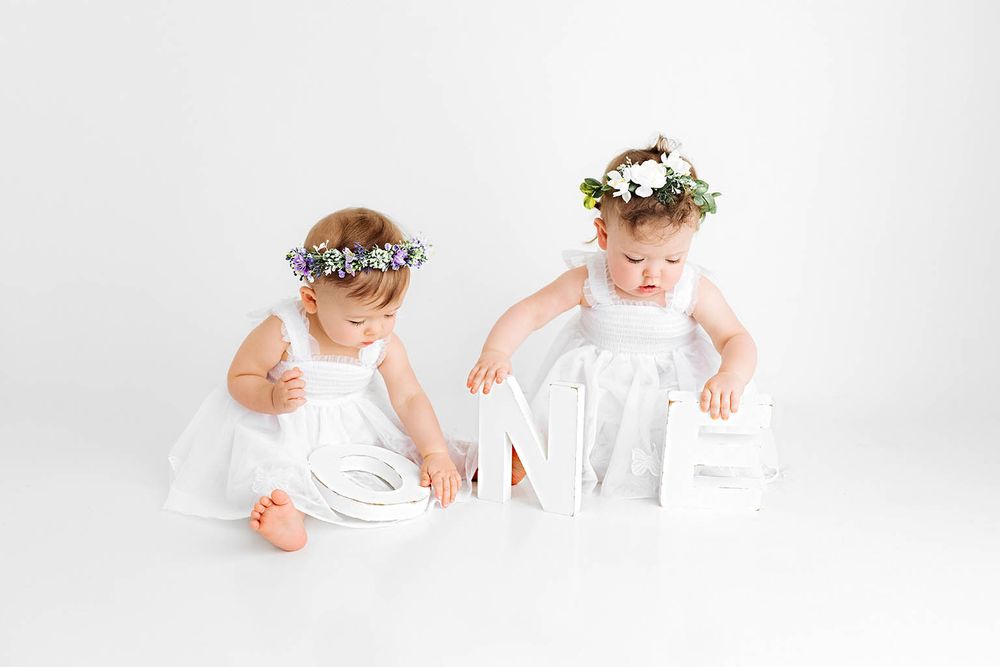 Two baby girls in white dresses and flower crowns playing with white decorative letters.