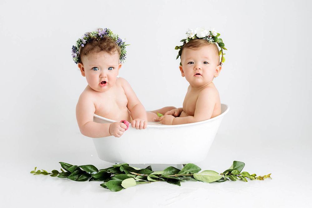 Two babies wearing flower crowns sitting together in a white bathtub with green leaves.